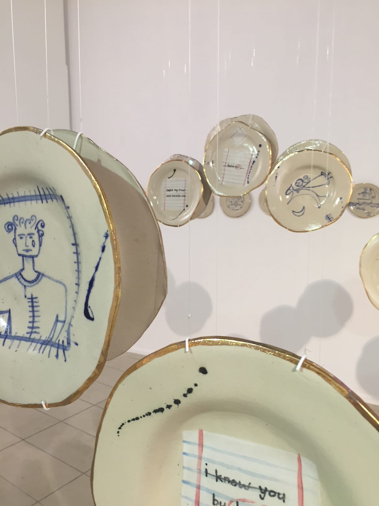 Ceramic plates with gold-edged rims hang suspended by strings. Several plates display hand-drawn blue sketches, such as a person and writing, whilst others have scribbled lines or abstract designs. The background is a white gallery wall and tiled floor.