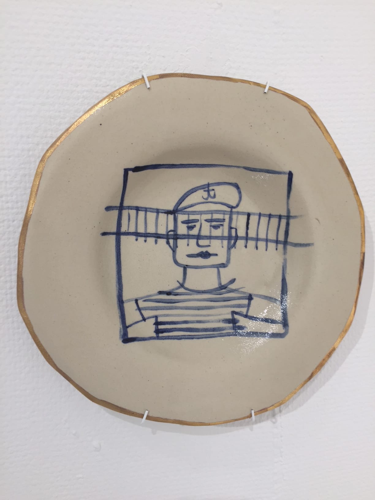 A beige ceramic plate with a gold-rimmed, uneven edge features a blue line drawing of a man wearing a beret and striped shirt, enclosed in a square with horizontal lines behind him. The plate is mounted on a white textured wall.
