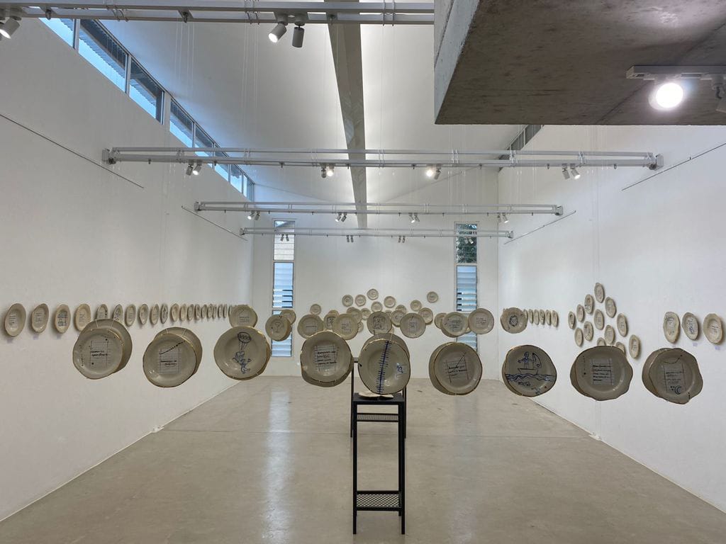 Art installation in a white gallery: numerous ceramic plates with drawings and text hang suspended in mid-air at varying heights, forming rows along the length of the room. Natural and artificial light illuminate the clean, minimalist space.